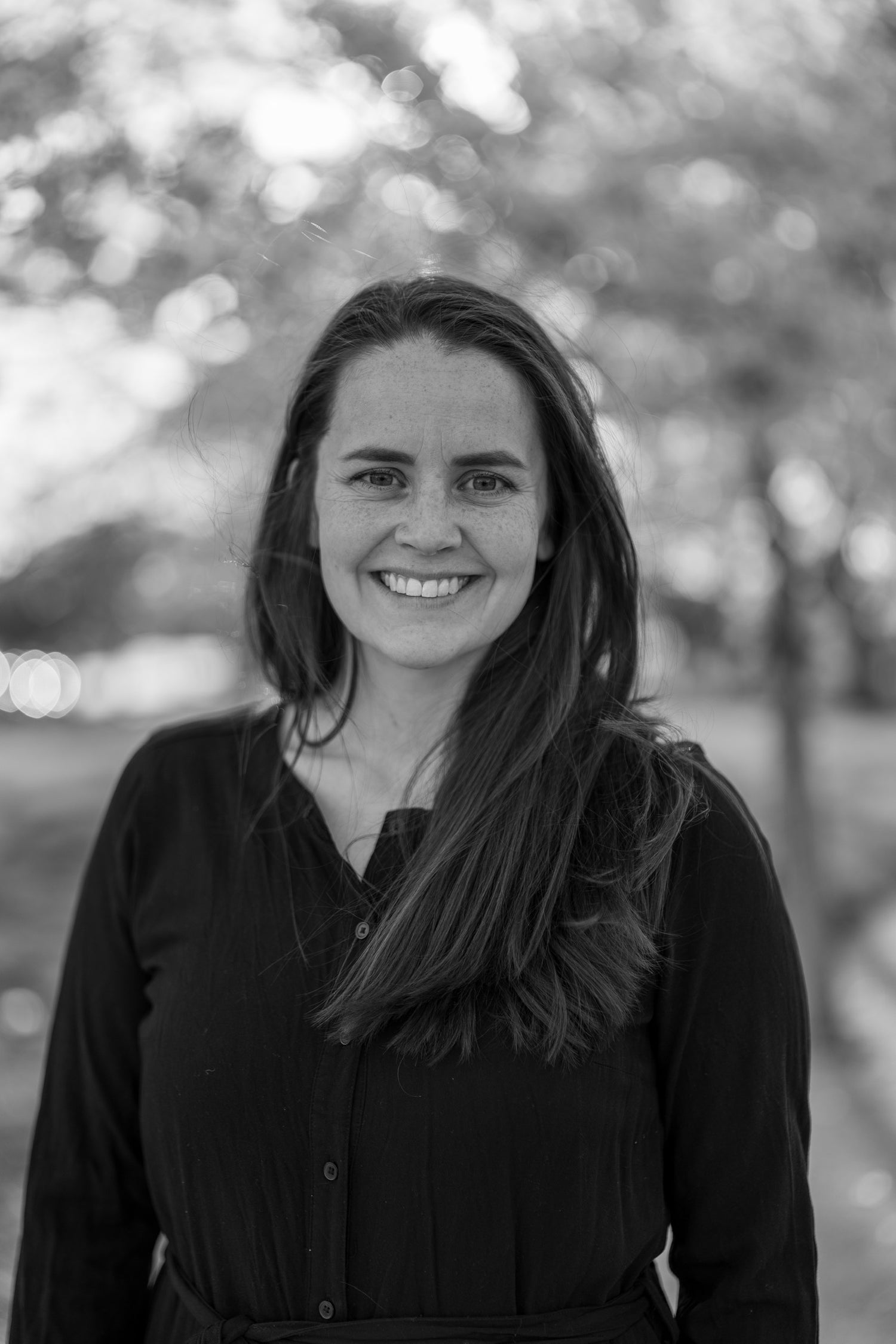 Jacqueline Christensen, the owner and designer of Jaykaiy Patterns. Black and white photo of Jacqueline with cherry blossoms in the background. Jacqueline is wearing a black jumpsuit.