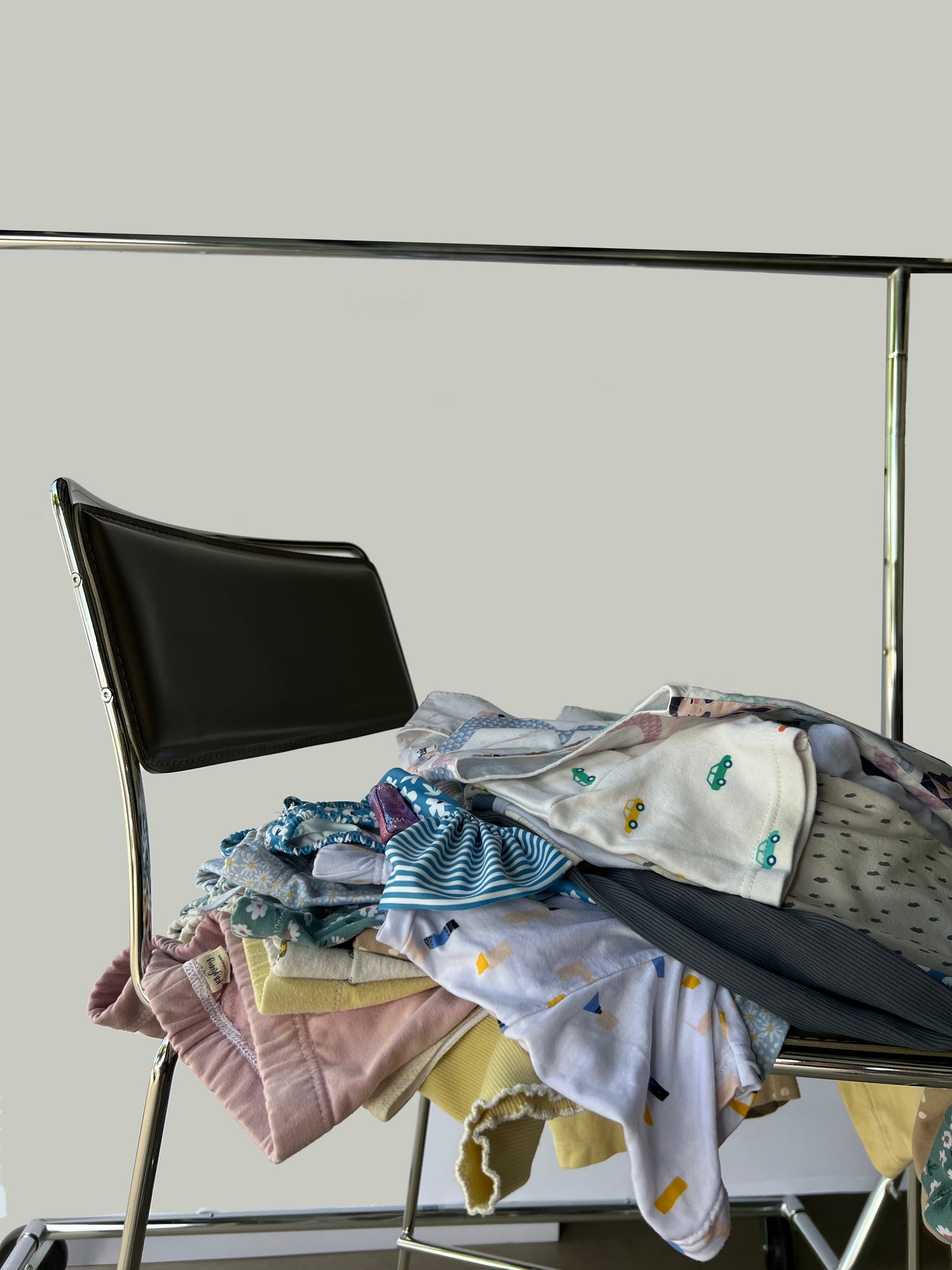 A pile of children's clothing layin on top of a tall chair with black back rest. In the background is a faded white background and clothing rack.