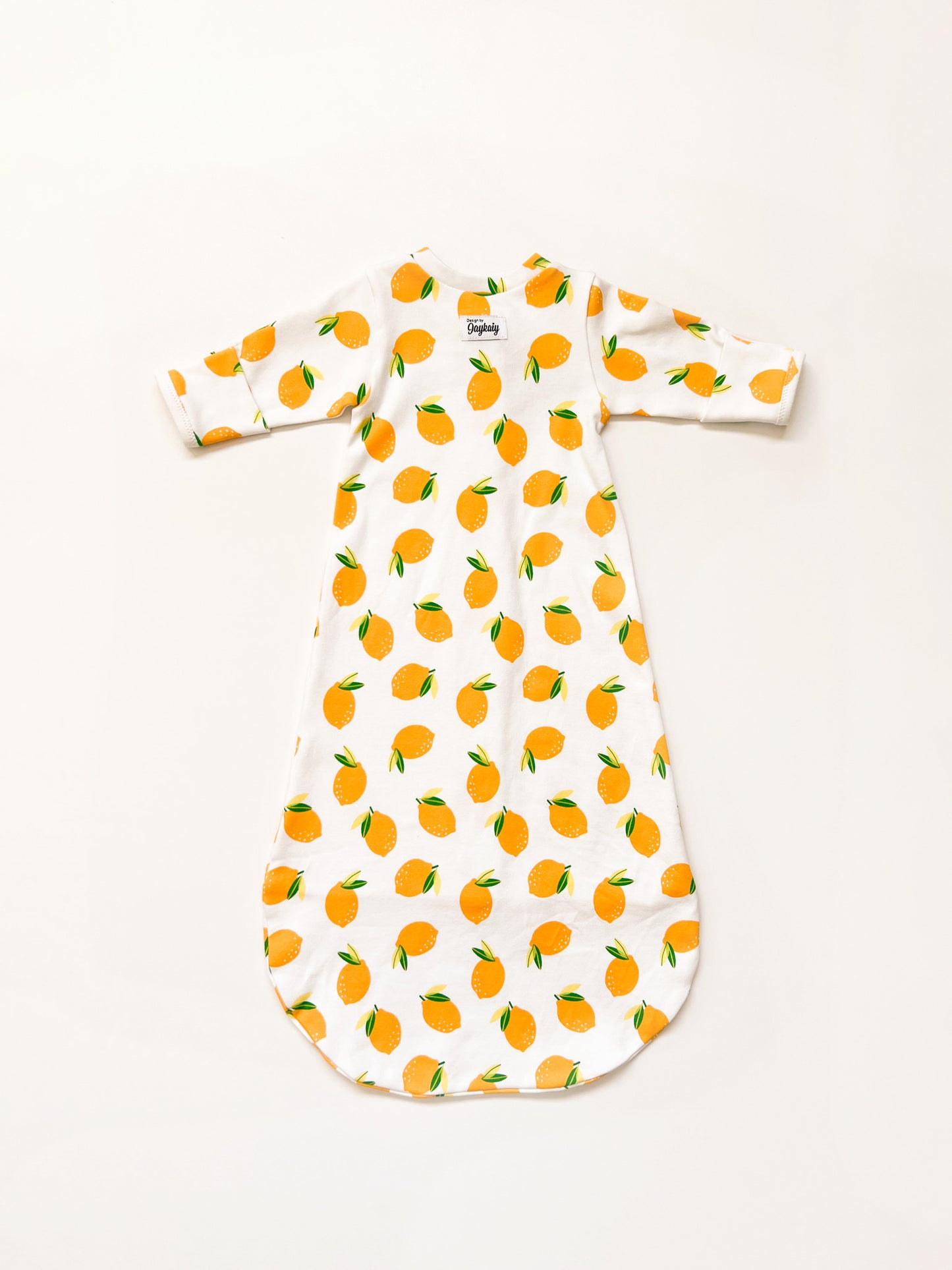 Flatlay, backside, of a baby sleepsack with zipper. The sleepsack is made in a white knit fabric with lemon print. Visible at upper back, centered below the neckline, is a Design by Jaykaiy tag.